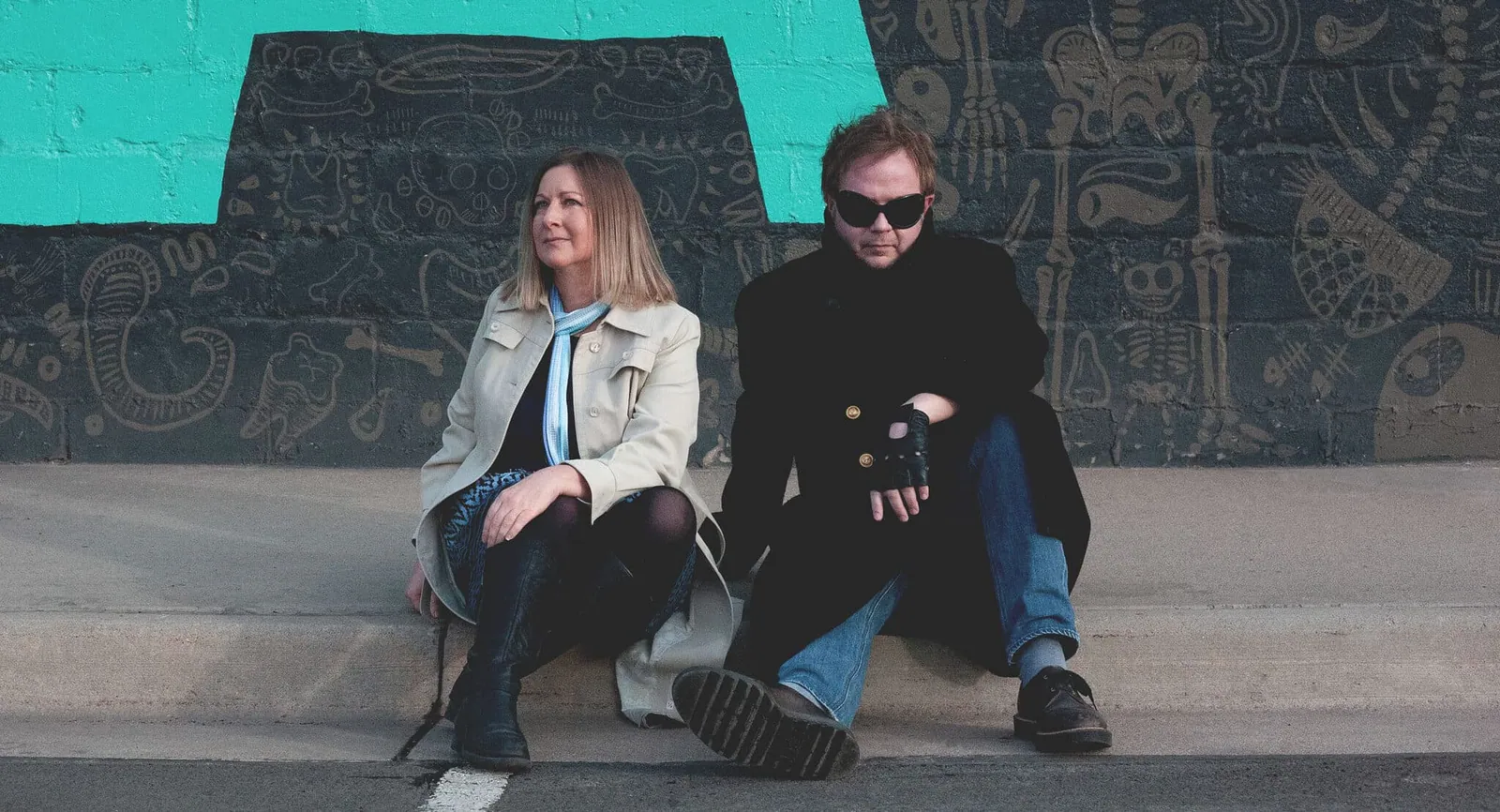 A woman in a cream jacket and a man in sunglasses sit on a concrete ledge before a dark, painted mural.