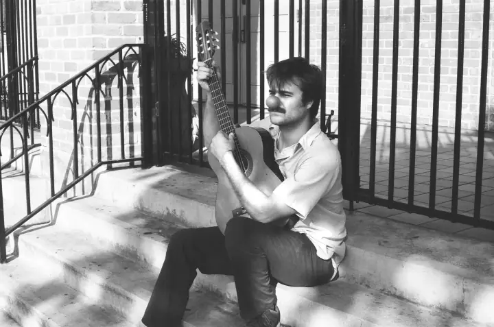 Black-and-white photo of a mustached man playing an acoustic guitar on concrete steps beside iron railings.
