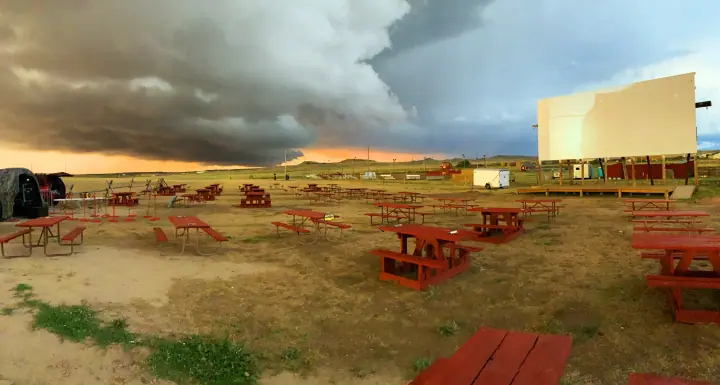 Empty red picnic tables face a large outdoor movie screen under a dramatic storm sky at sunset.