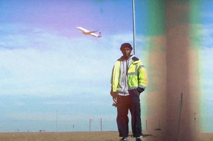 A man in a neon yellow high-visibility jacket and dark beanie stands on dirt as a plane flies overhead.