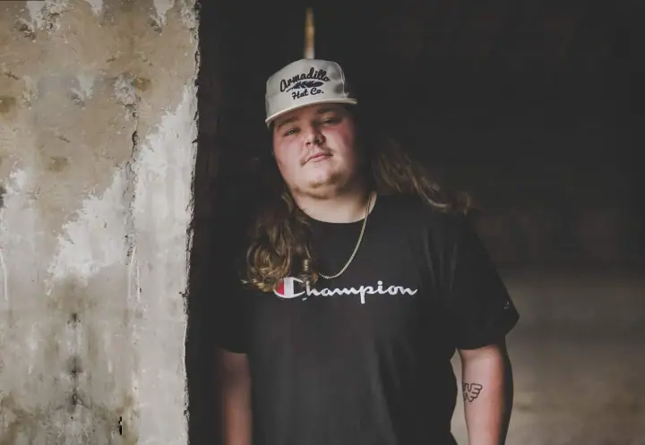 Long-haired man in a gray cap and black Champion tee leans against a weathered concrete wall indoors.