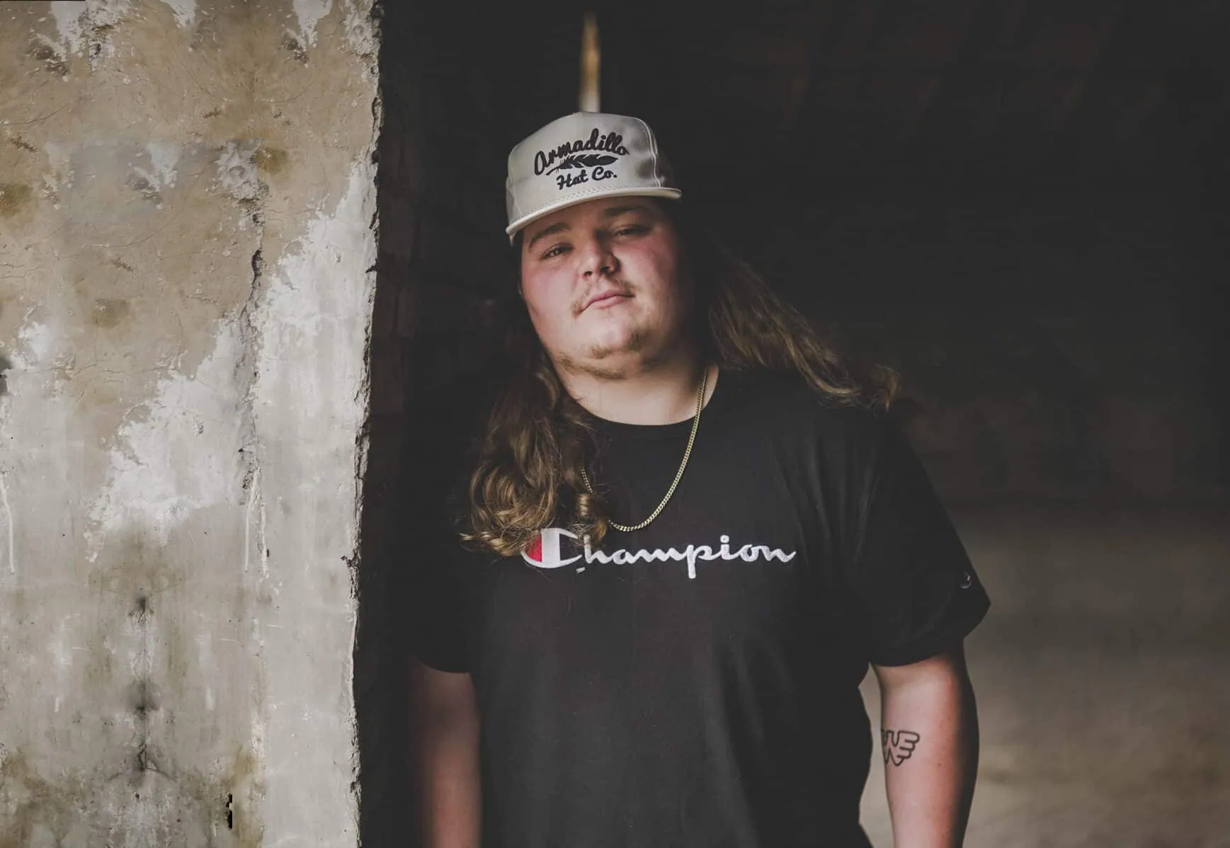 Long-haired man in a gray cap and black Champion tee leans against a weathered concrete wall indoors.