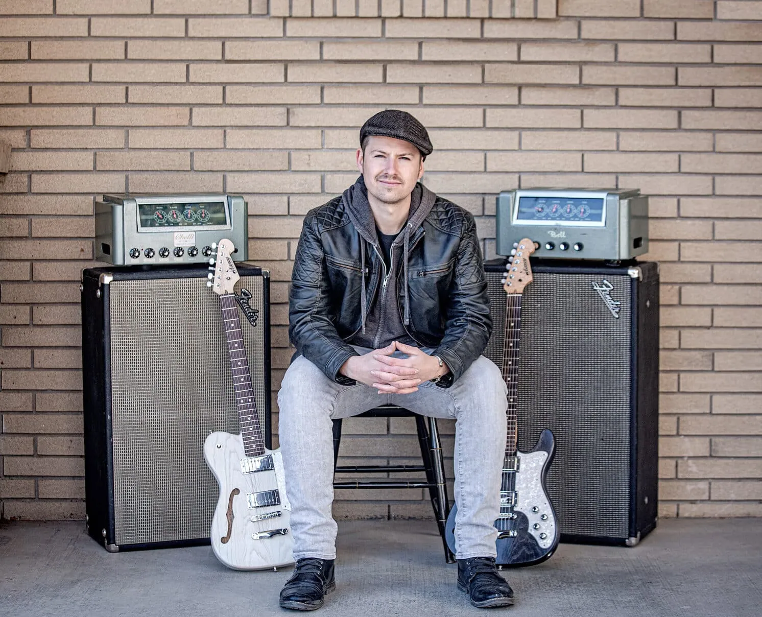 A man in a leather jacket and flat cap sits between two guitar amps with electric guitars against a brick wall.