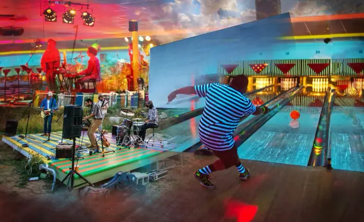 Band performs on a striped stage while a person in a striped shirt bowls in a colorful venue.