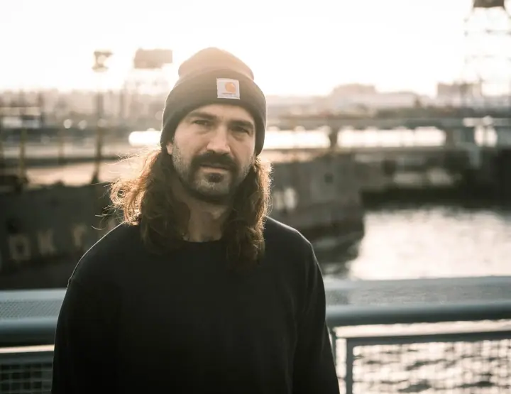 A bearded man with long brown hair and a black beanie stands by a railing near a waterfront in warm light.