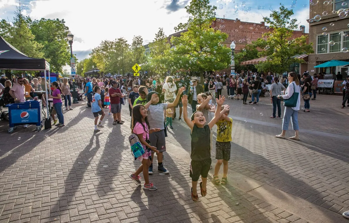 Children reach for floating bubbles on a brick-paved street crowded with festival crowds, tents, and a Pepsi cooler.