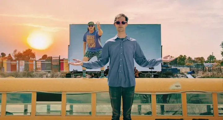 A man in a blue shirt and red sunglasses spreads his arms before a sunset billboard reading MAKE MORE FILMS.