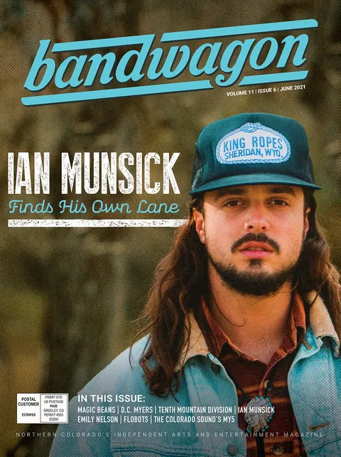 Ian Munsick wears a teal King Ropes trucker cap, denim jacket, and bolo tie outdoors in a blurred green setting.