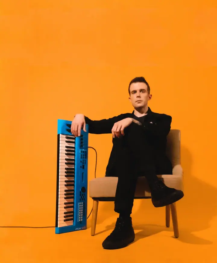 A man in black sits in a beige armchair beside a blue MX49 keyboard against a bright orange backdrop.