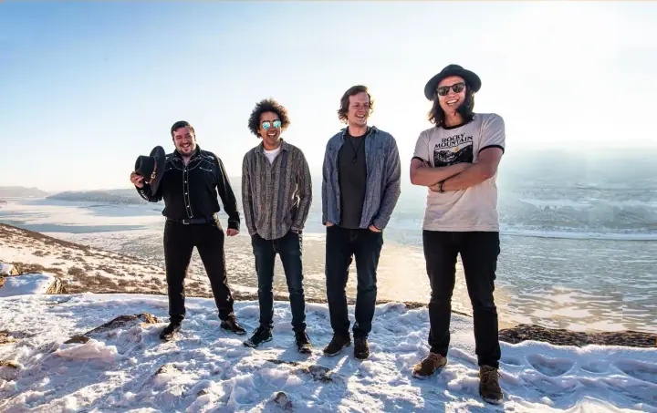 Four men smile on a snowy hilltop above a frozen lake, one holding a black hat and another wearing sunglasses.