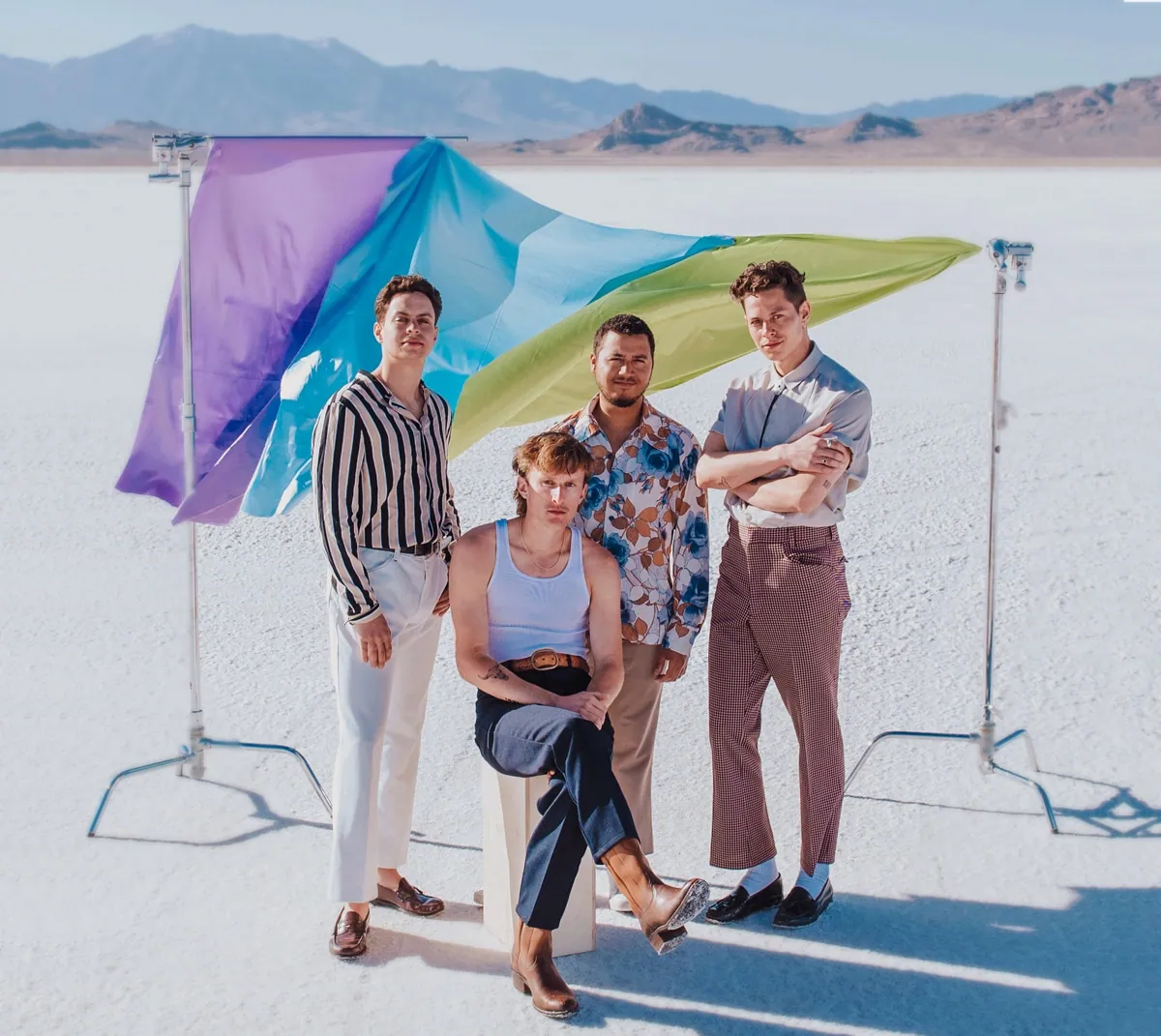 Four men pose on a white salt flat in front of a flowing purple, blue, and green flag with mountains behind them.