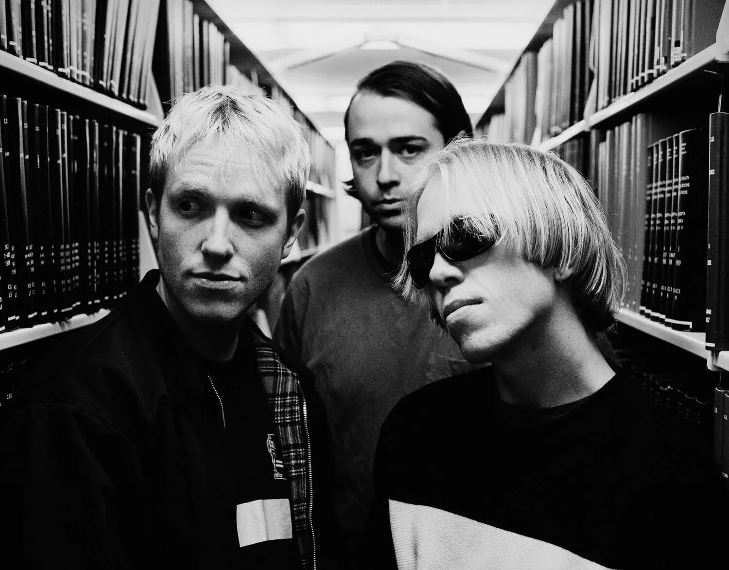 Black and white portrait of three men posing between library bookshelves, one wearing sunglasses.