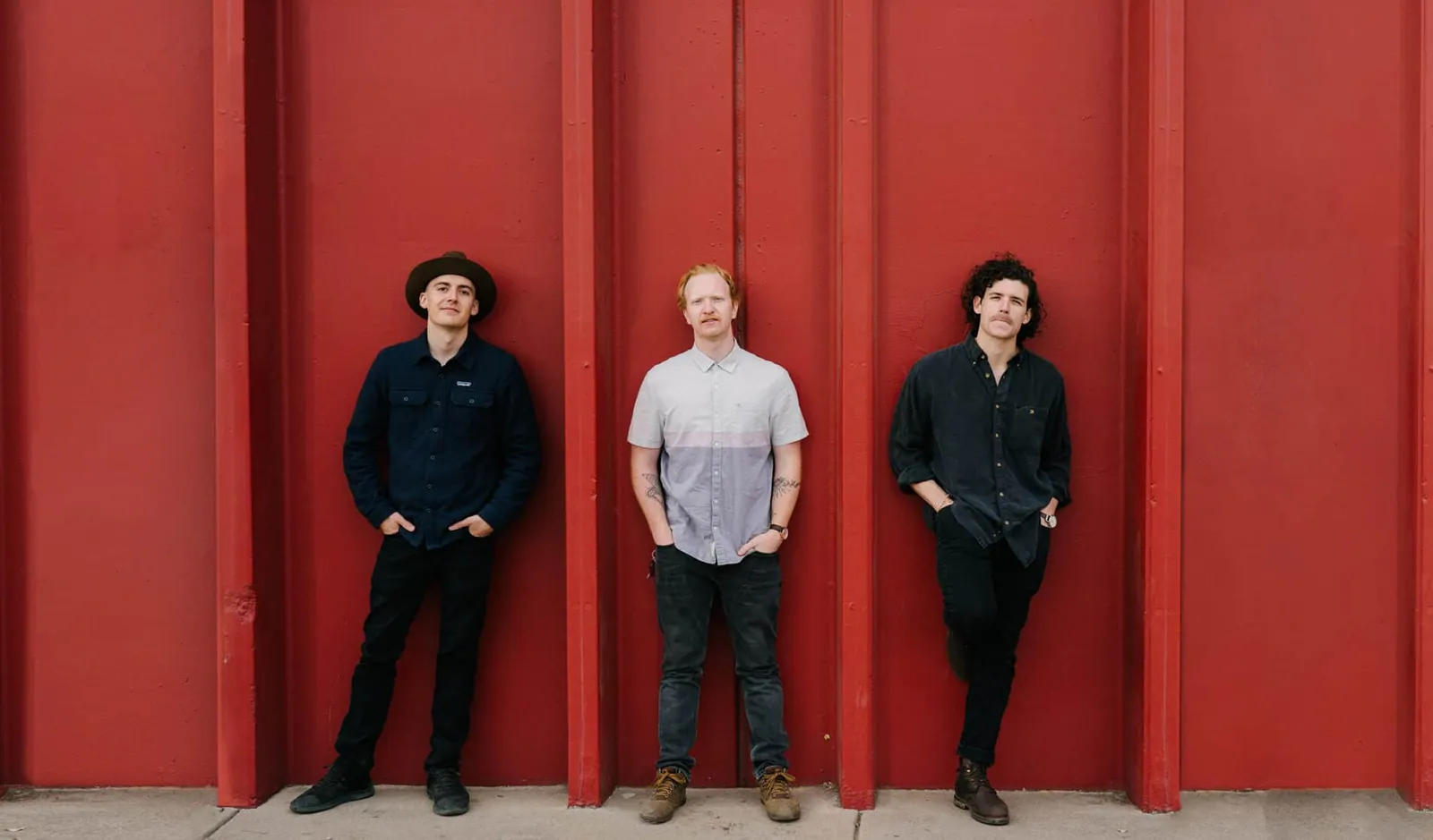 Three men in dark and gray shirts stand against a red metal wall, hands in pockets, with no instruments visible.