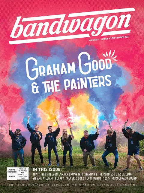 Seven men in matching black shirts hold colored smoke flares in a grassy field under a pink sky.