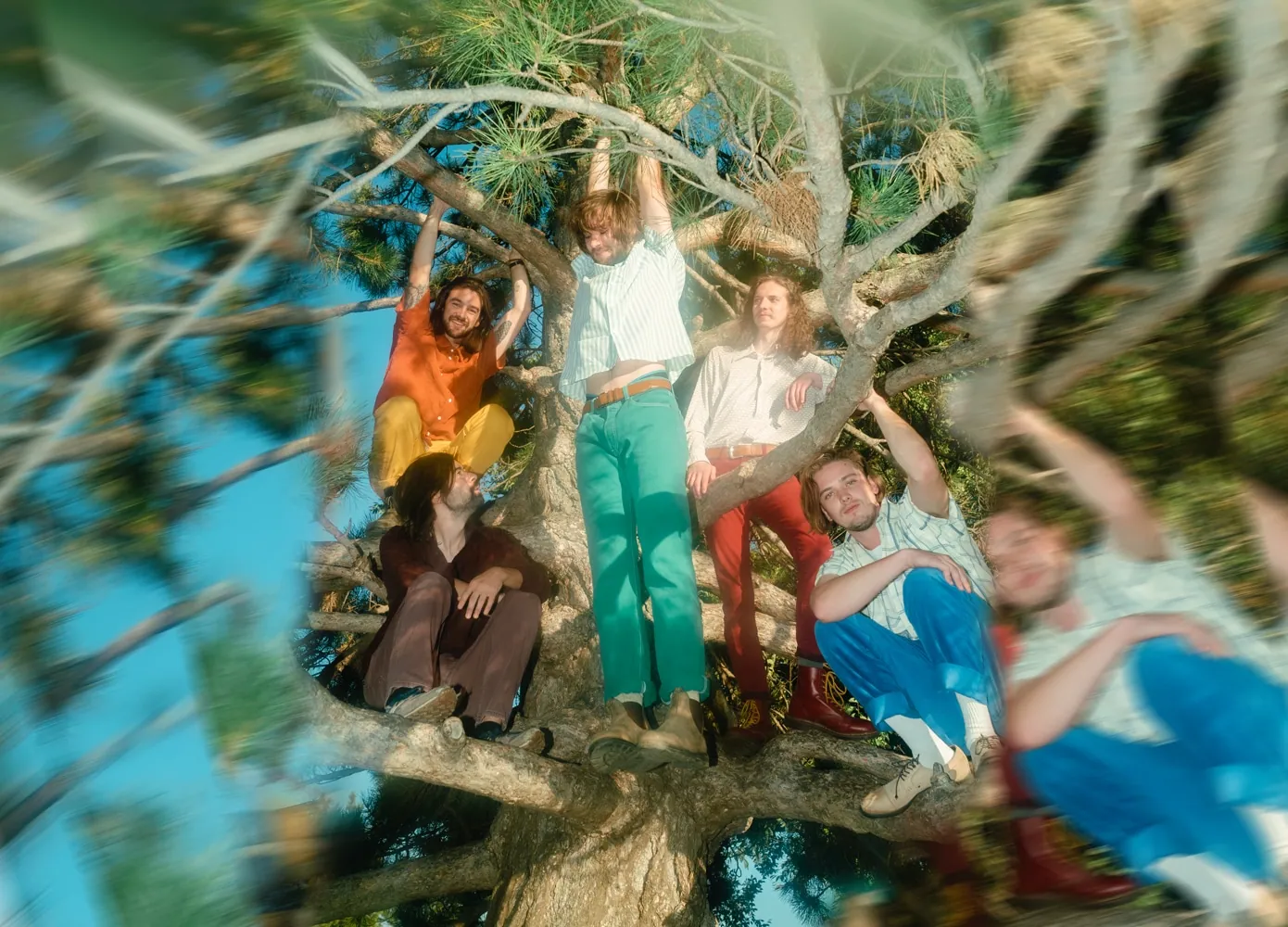 Six people in bright vintage clothing lounge and climb through a tree with blurred branches against a blue sky.