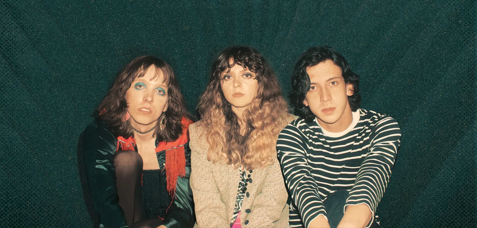 The Velveteers: three members pose against a dark green backdrop, one in a satin top with red fringe.