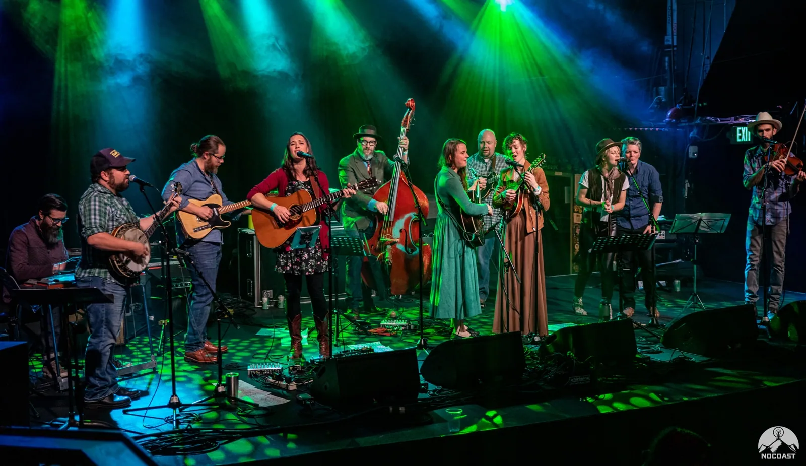 A large folk ensemble performs under green and blue stage lights, with banjo, guitars, upright bass, fiddle, and keys.