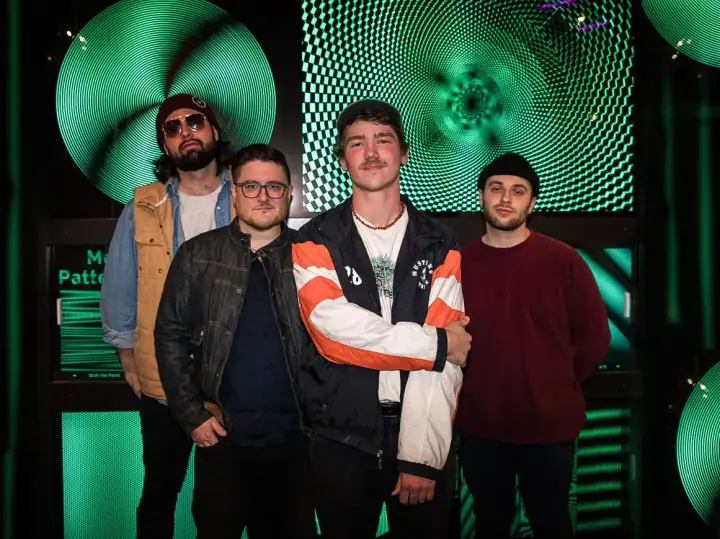 Four men pose together in front of glowing green circular light displays at a venue, with no instruments visible.