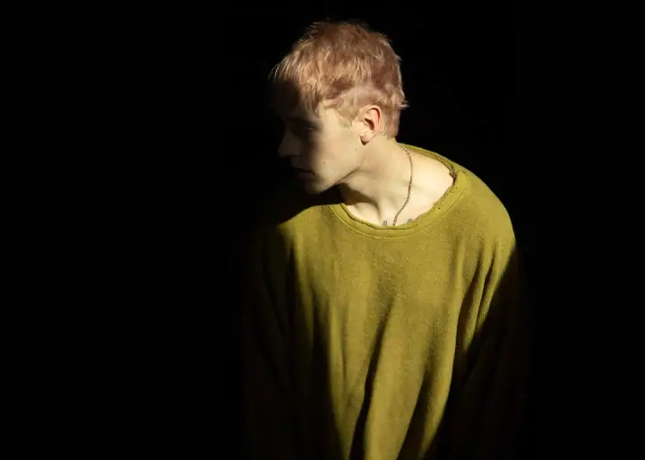 Person with short pink-blond hair in an olive-green sweater and chain necklace, lit against a black background.