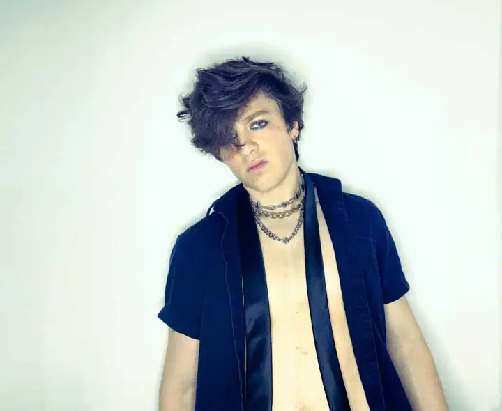 Young person with tousled dark hair and layered chains wears an open navy shirt against a plain white backdrop.