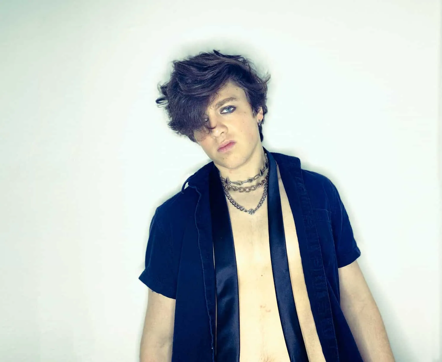 Young person with tousled dark hair and layered chains wears an open navy shirt against a plain white backdrop.