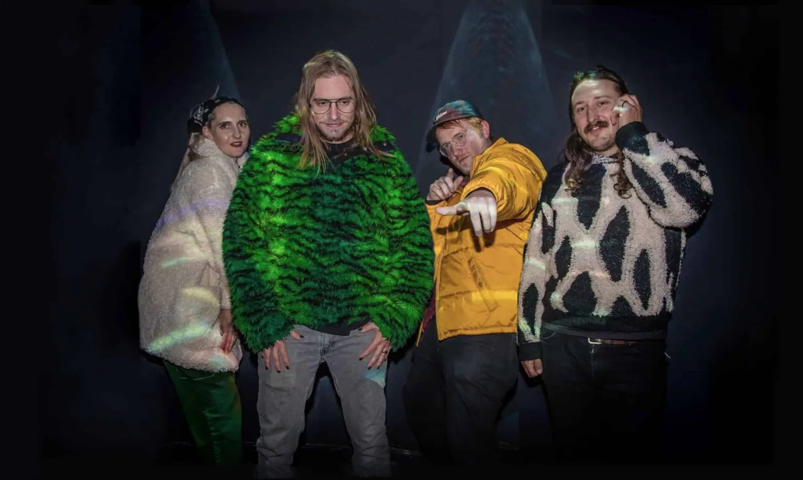 Four people in fur and patterned knitwear pose in a dark room lit by spotlight beams.