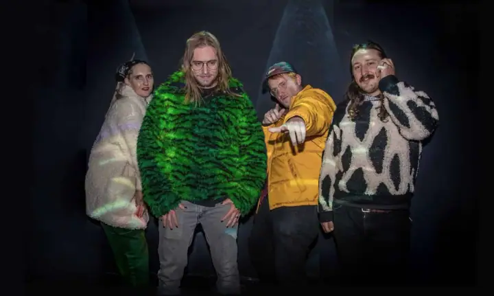 Four people in fur and patterned knitwear pose in a dark room lit by spotlight beams.