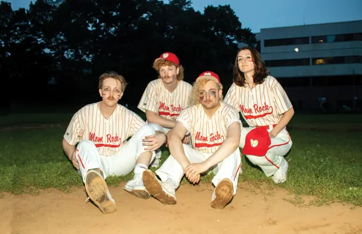 Mom Rock members in pinstriped baseball jerseys and red caps pose on a dirt infield at dusk.