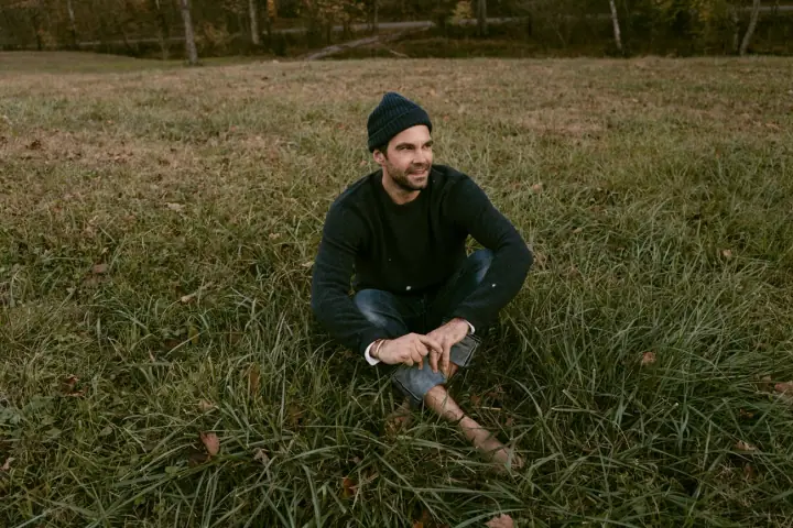 A man in a dark knit beanie and sweater sits cross-legged in a grassy field, smiling and gazing to the side.