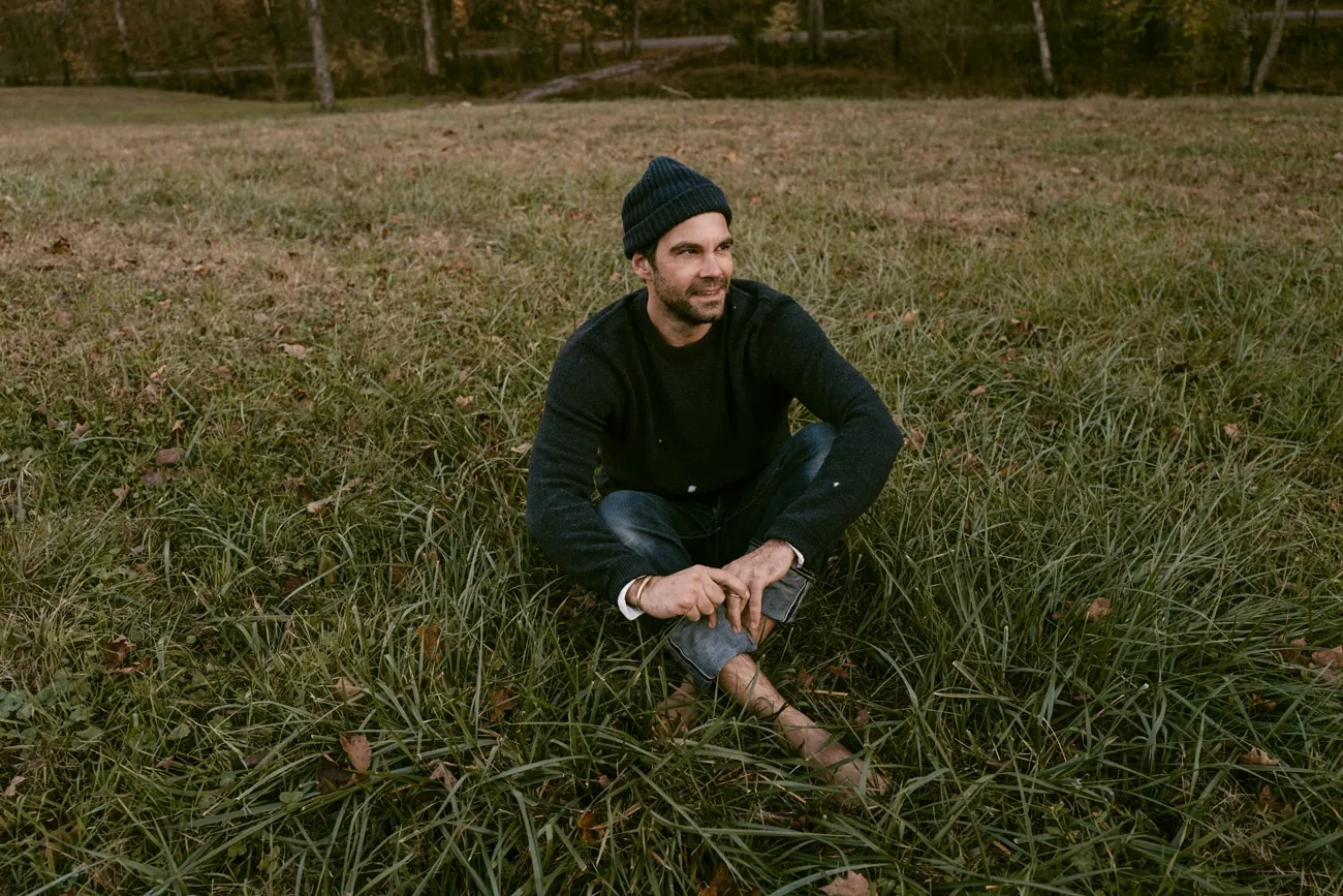 A man in a dark knit beanie and sweater sits cross-legged in a grassy field, smiling and gazing to the side.
