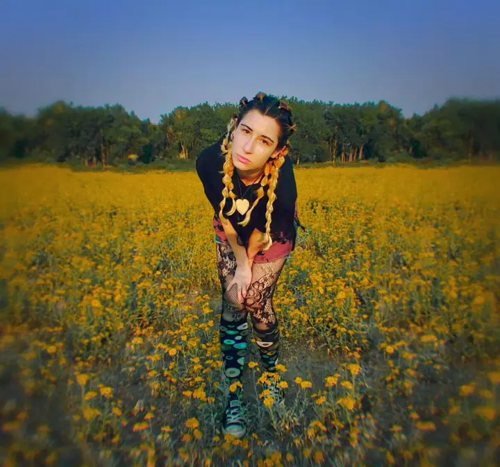 Person with blonde braids and a black top leans forward in a yellow wildflower field, wearing fishnets and patterned socks.