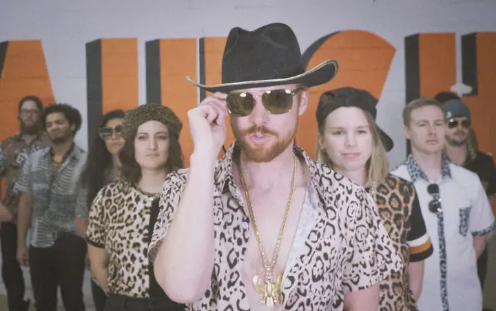 A bearded man in a cowboy hat and leopard shirt leads a group in front of an orange and black wall.