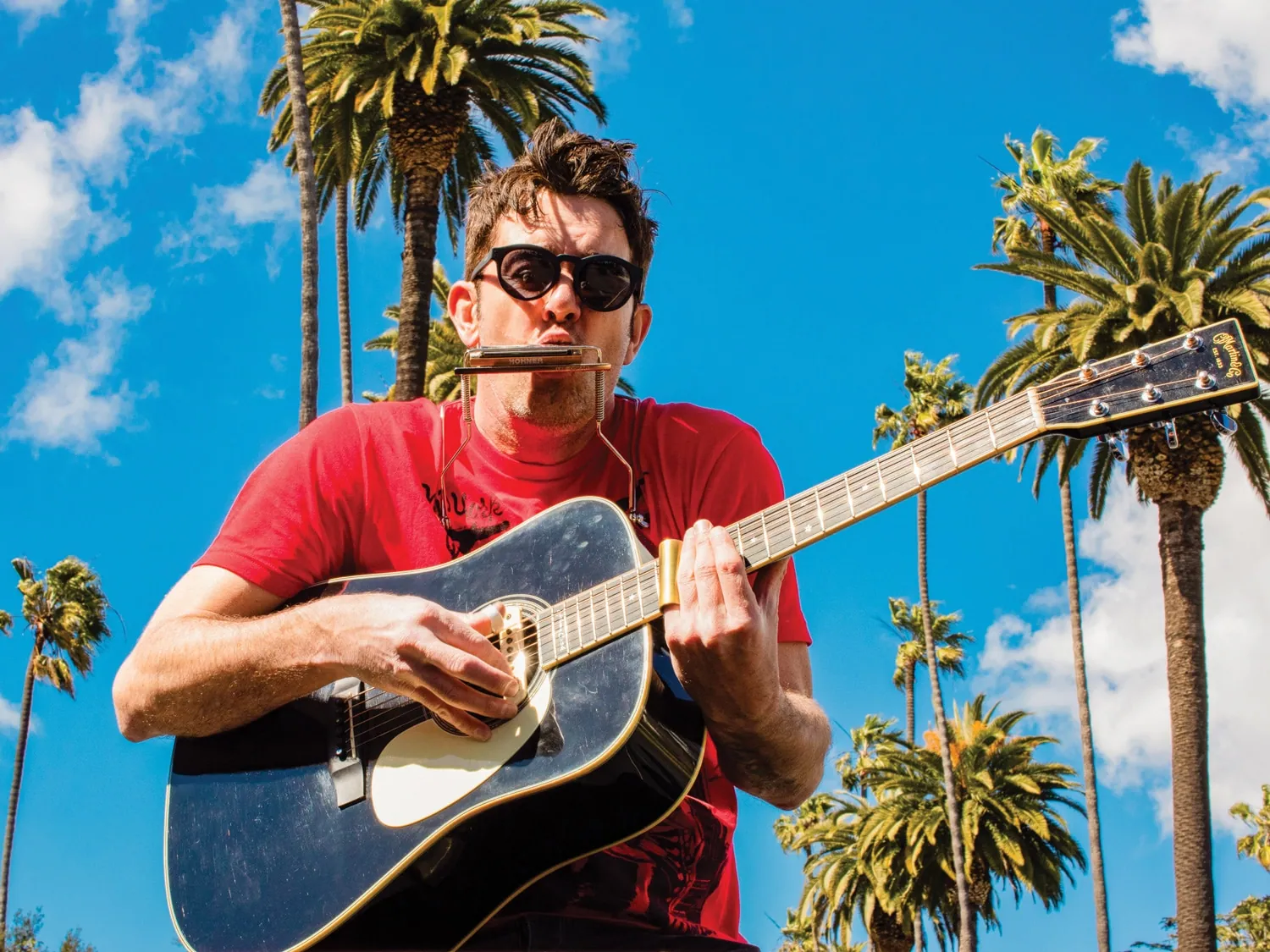 A man in sunglasses and a red t-shirt plays an acoustic guitar and harmonica under tall palm trees and a blue sky.