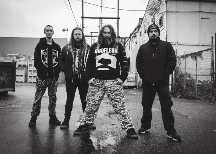 Four men in jackets and camouflage pants stand on a wet alley pavement beside a chain-link fence in black and white.