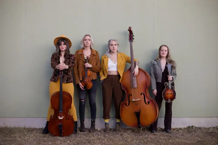 Four women stand against a sage green wall holding a cello, violin, upright bass, and mandolin outdoors.