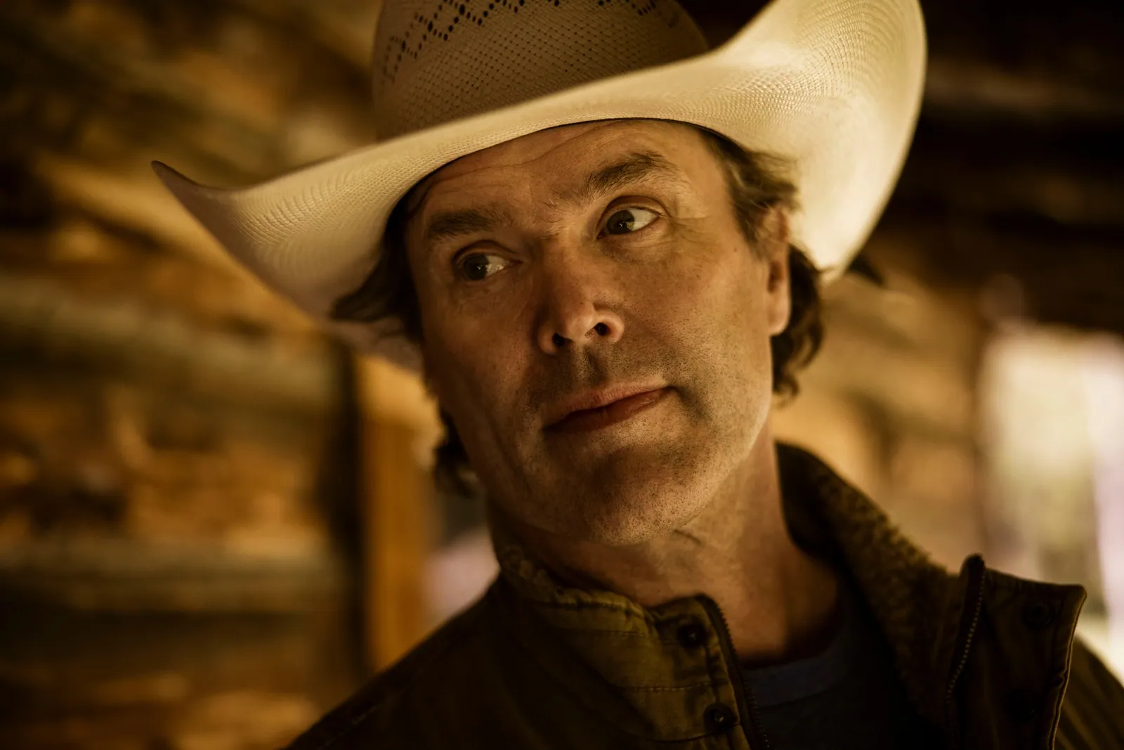 Man in a cream woven cowboy hat and dark jacket glances sideways in a dimly lit room with rustic log walls.