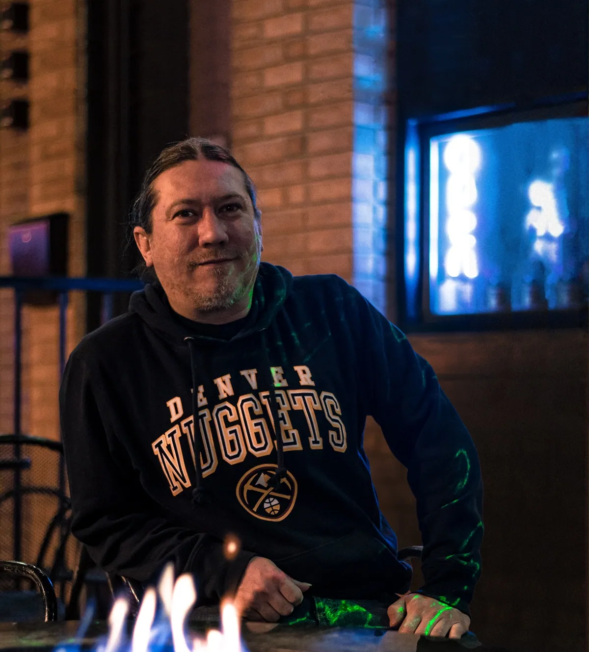 A smiling man with his hair pulled back wears a black Denver Nuggets hoodie at a bar, with a candle flame in front.