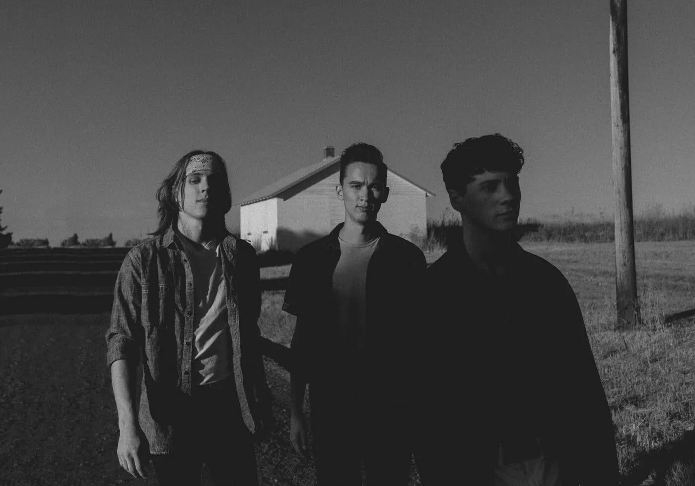 Black and white photo of three young men standing in a field before a white farmhouse and utility pole.
