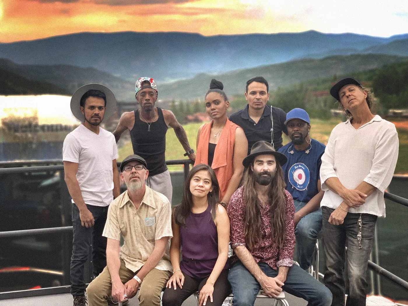 Nine people pose on a railed deck before a sunset mountain backdrop, with no instruments visible.