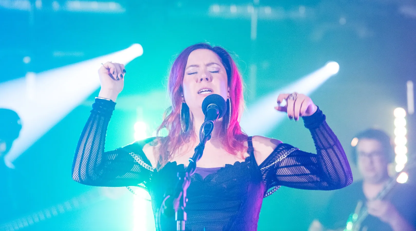 Kaitlyn Williams sings into a mic with raised arms under teal stage lights, a blurred guitarist behind her.