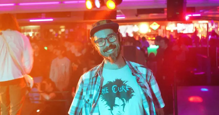 A smiling bearded man in a cap, glasses, and plaid shirt stands in a crowded club under pink and purple lights.