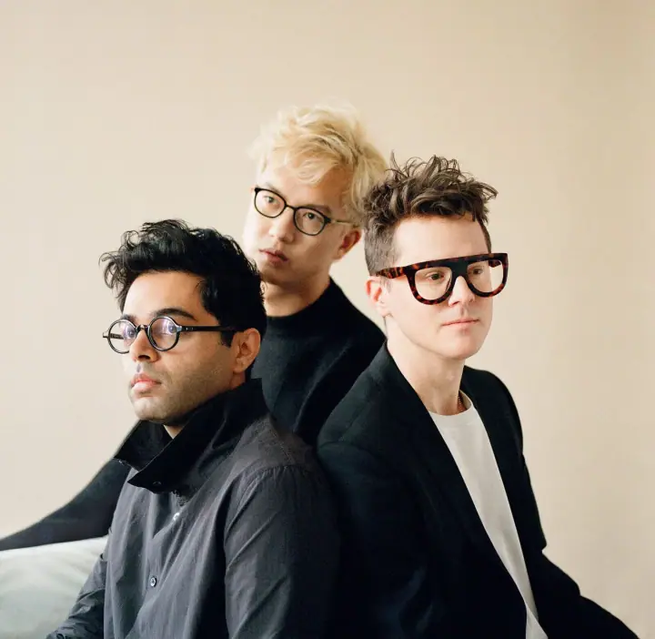 Son Lux band portrait: three people in dark clothing and glasses posed against a plain cream wall.