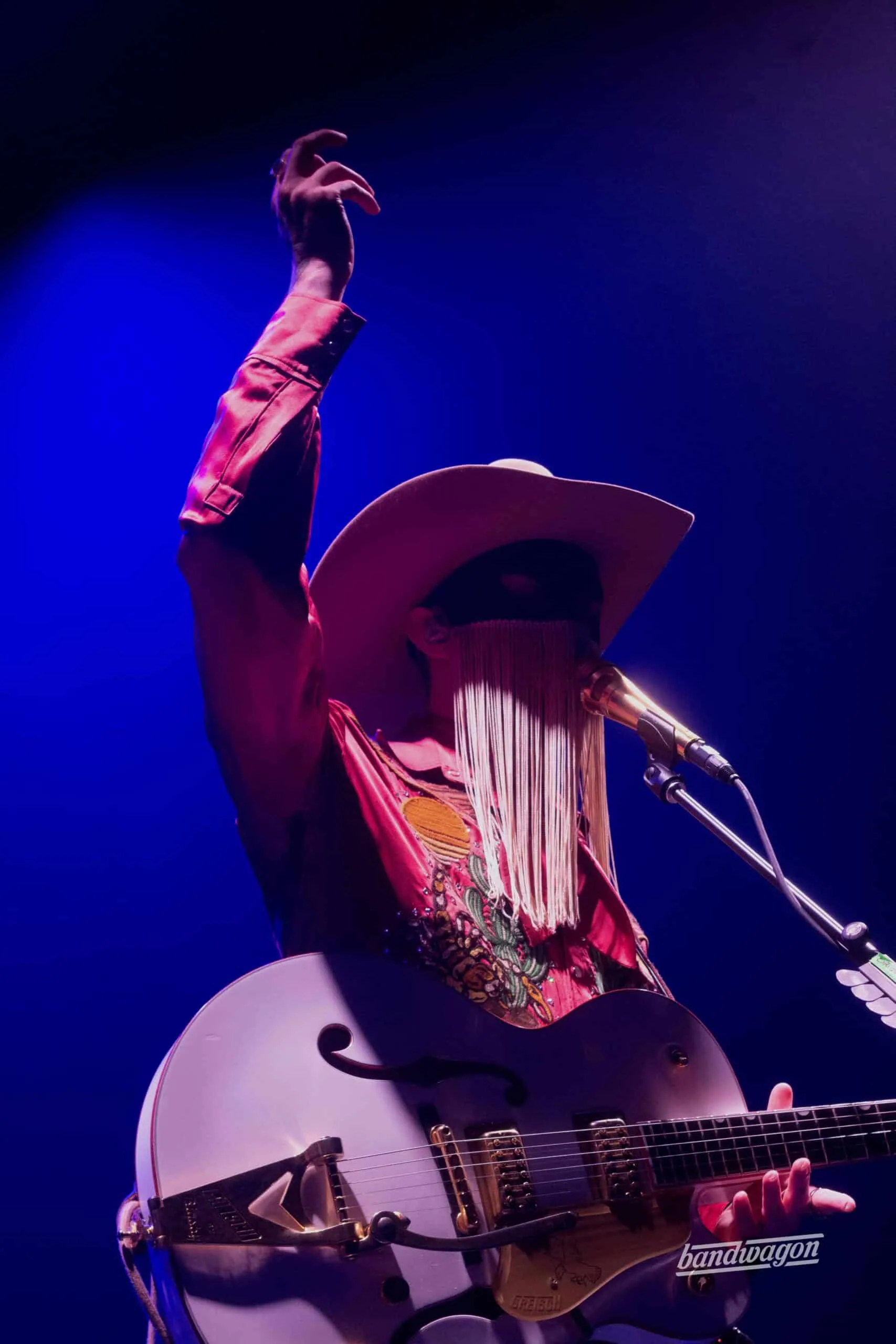 Orville Peck raises one arm on stage in a fringed mask and cowboy hat while playing a white guitar.