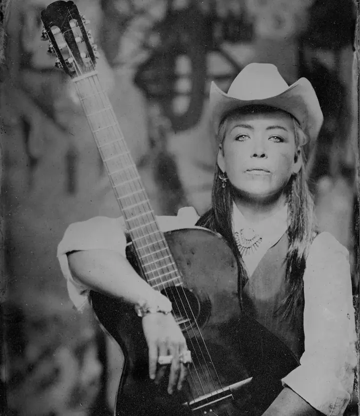 Black-and-white portrait of a woman in a cowboy hat holding an acoustic guitar against a blurred backdrop.