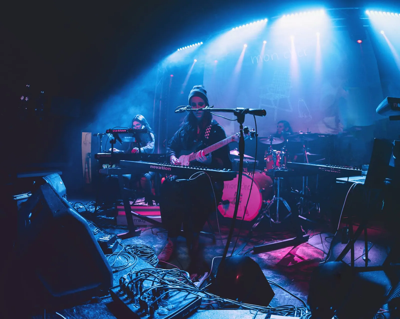 Band performs under blue stage lights, with a guitarist, keyboardist, and drummer behind a red drum kit.