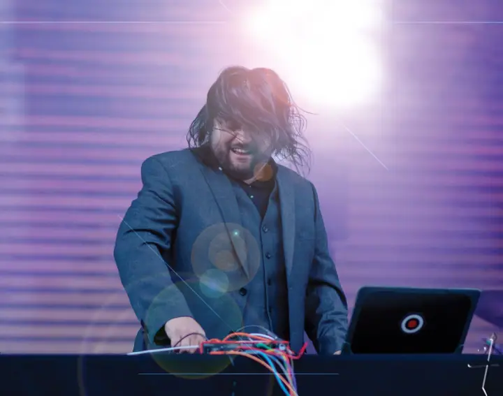 A man with long dark hair and a gray blazer plays a mixing board with colorful cables under purple stage lights.