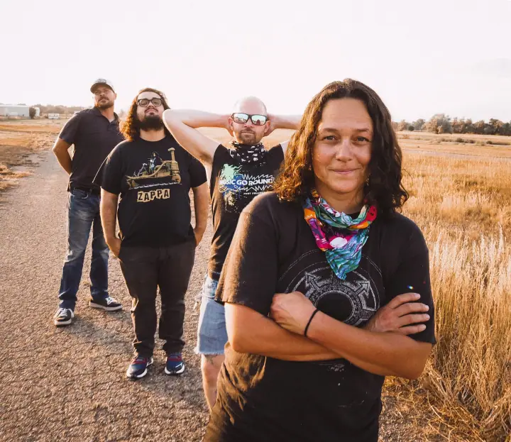 Four people stand on a gravel road in dry grass, the woman in front with arms crossed and a bright bandana.