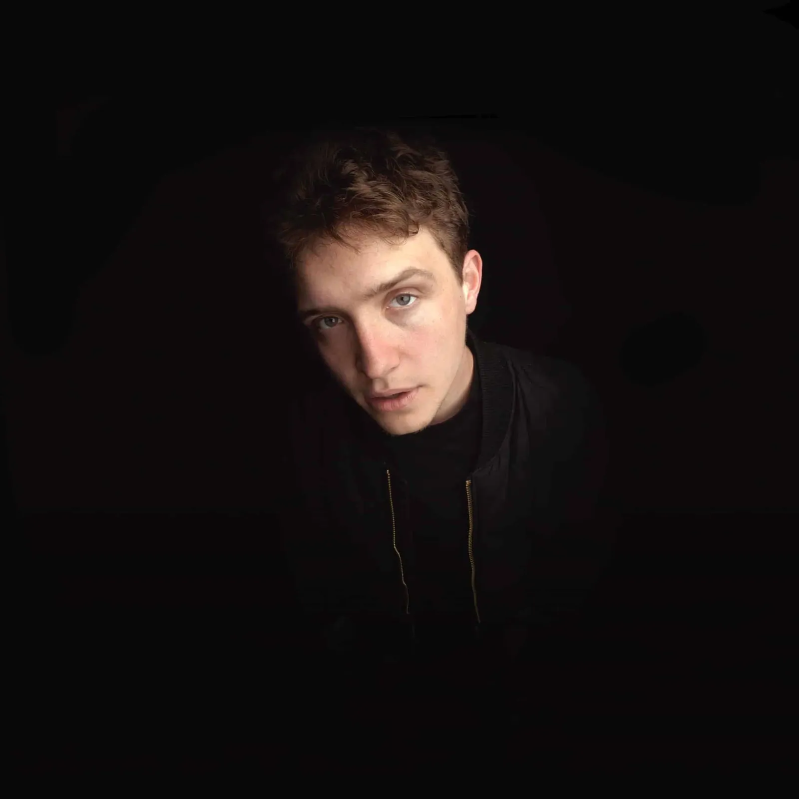 A young man with tousled brown hair and a dark zip-up jacket gazes at the camera against a black background.