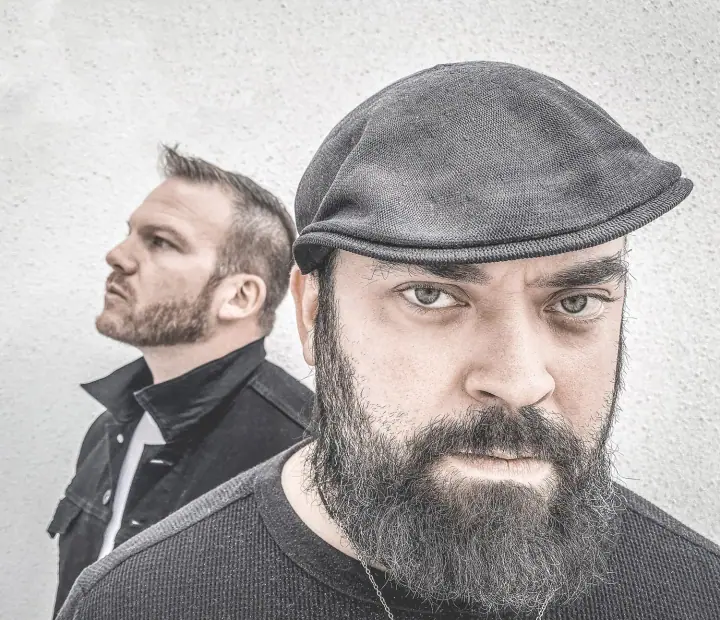 Two bearded men pose against a gray textured wall, one in a flat cap facing forward and one in profile behind him.