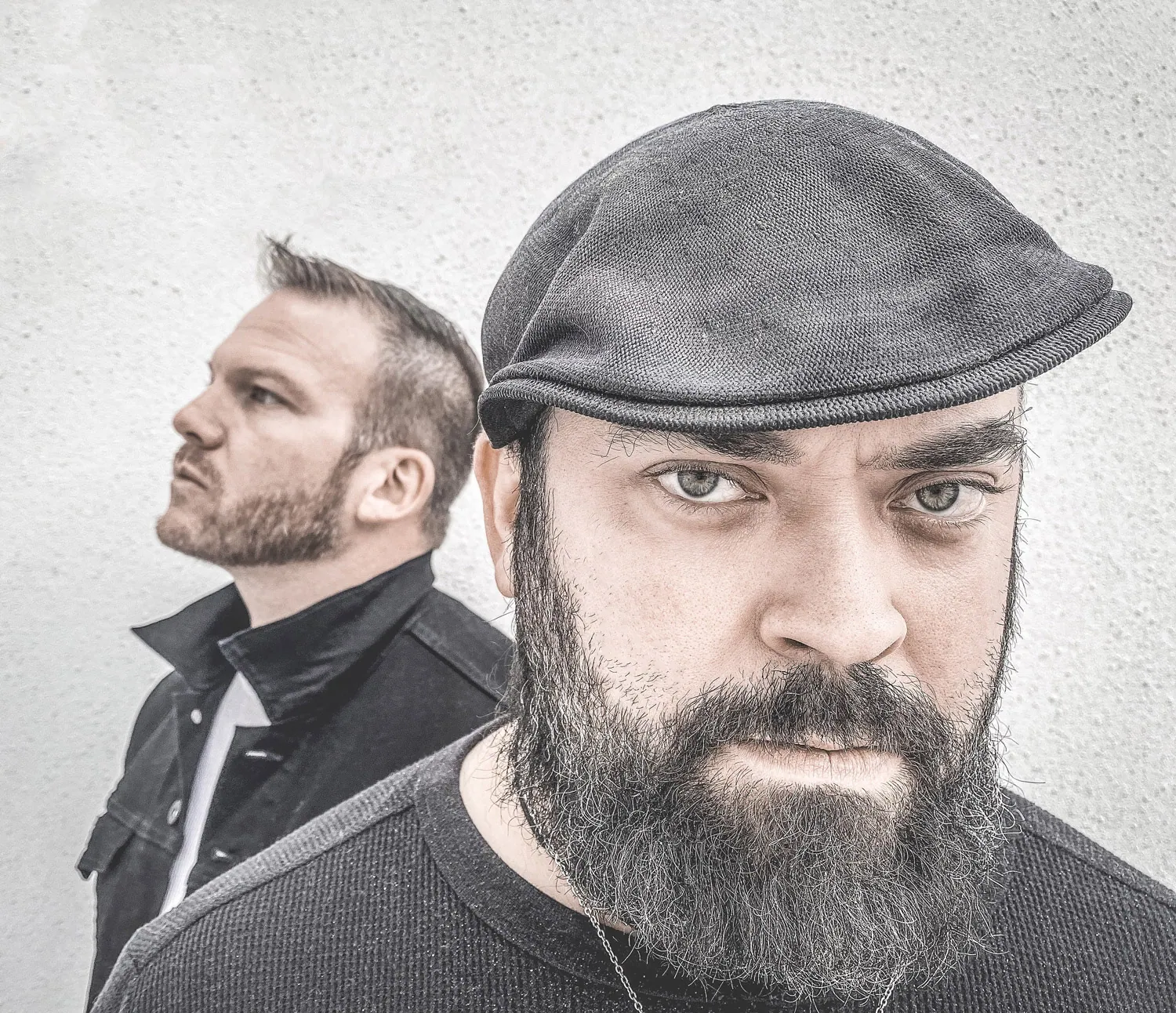 Two bearded men pose against a gray textured wall, one in a flat cap facing forward and one in profile behind him.